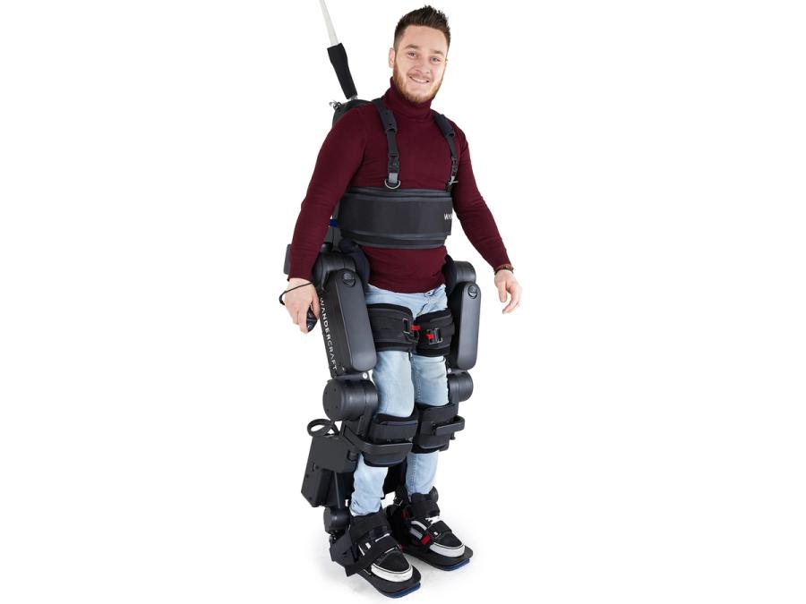 A smiling man in a dark red turtleneck and jeans in the Atalante X, a tethered exoskeleton that includes strapping around his legs and torso.