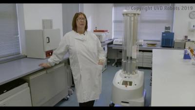 A woman in a white hospital coat stands next to a UV disinfection robot in a lab facility room.