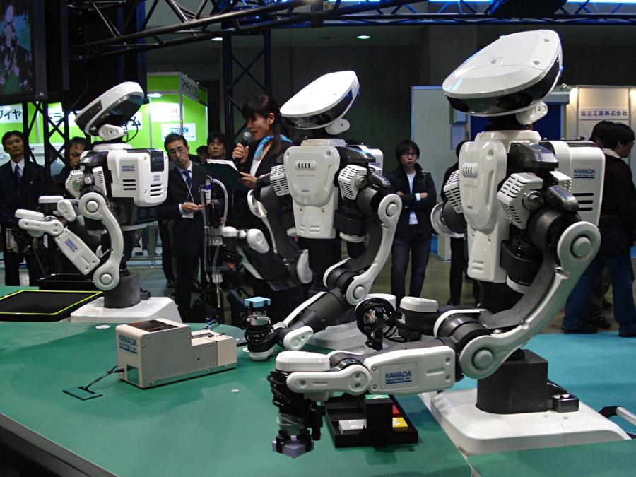 Three Nextage robots manipulate objects on a green surface at a trade fair.