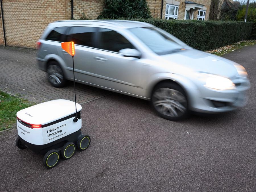 A white boxy mobile robot on six wheels with a narrow antenna with an orange flag pauses to let a moving car pass.