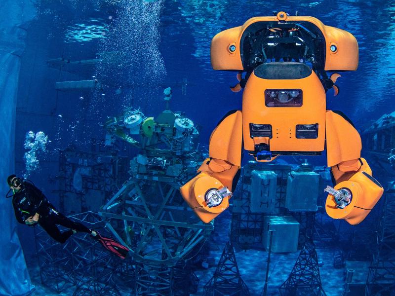 An orange half humanoid submarine robot with gripper hands is underwater in a space with a scuba diver.