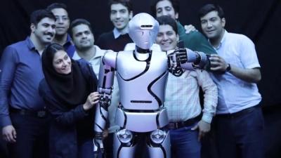 A group of people smile while standing behind a silver humanoid robot holding a smartphone to take a s
