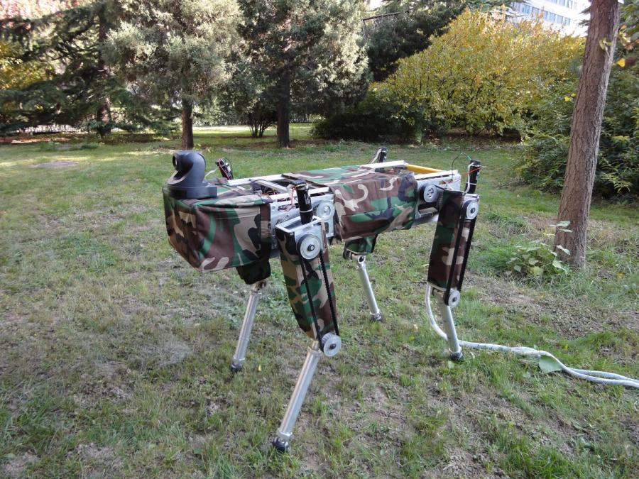 A quadruped robot stands on grass. It has a rectangular body and four legs, a black camera sitting on top, a tether, and is covered in camouflage fabric.