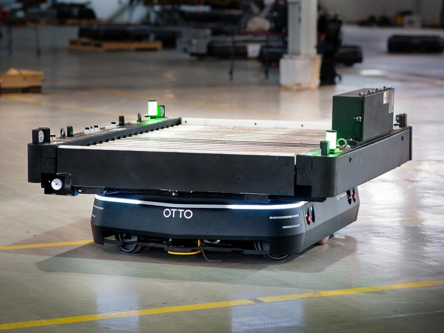 A squat, square, black and silver mobile robot carries a larger object across a factory floor.