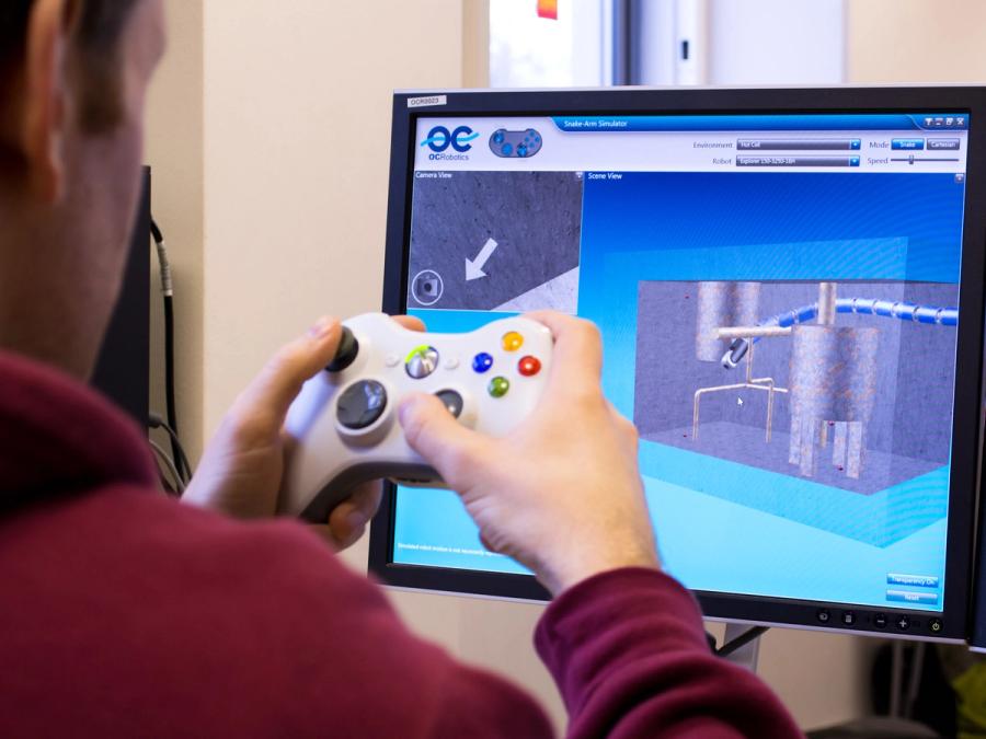 A person manipulates a game controller while looking at a monitor that shows the snake robot moving through an industrial space.