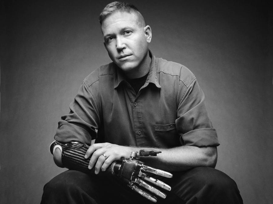 Black and white modern photograph of a seated man who sits with his flesh hand holding his robotic arm and hand.