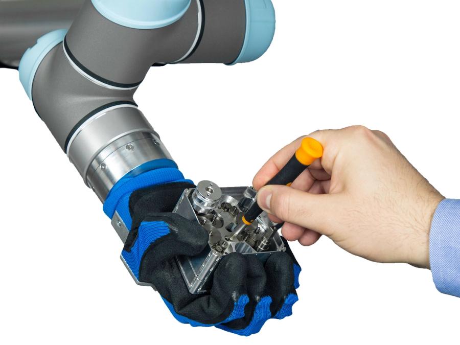 The hand is attached to a robotic arm. It grips a metal square, which a human hand is reaching into with a screwdriver.