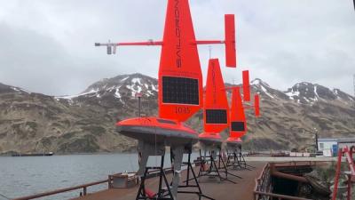 Three Saildrone robots, which look like small boats with red sails, await to be deployed from a large boat, with snowy mountains in the background.