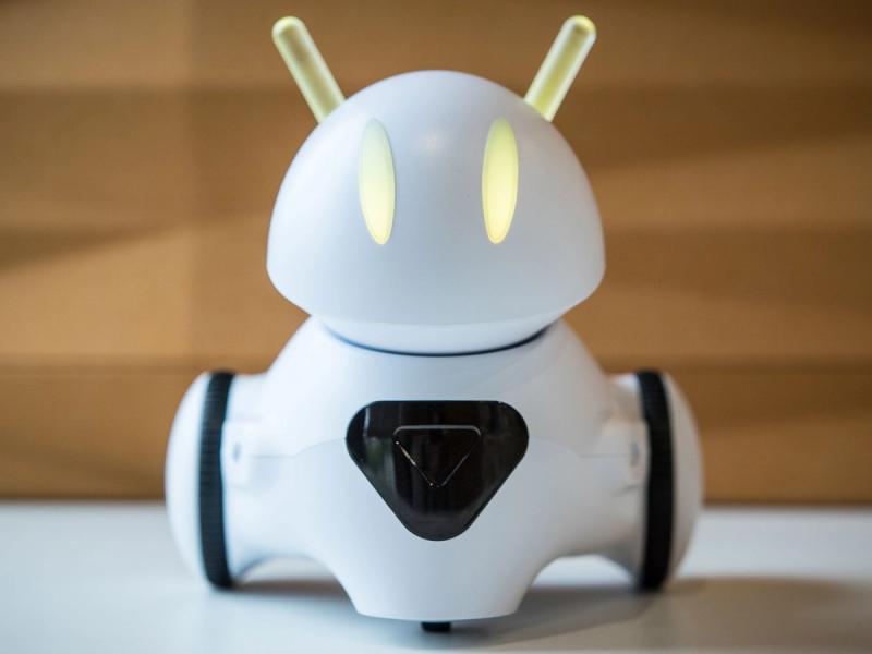 A simple, small, white robot with two wheels, a black camera in its chest, and yellow glowing eyes and antenna ears.