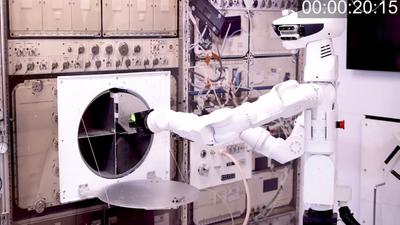 Space robot GITAI G1, a humanoid with a white metal body with a sensor-packed head and articulated arms, manipulates a pieced of equipment in a simulate space station.