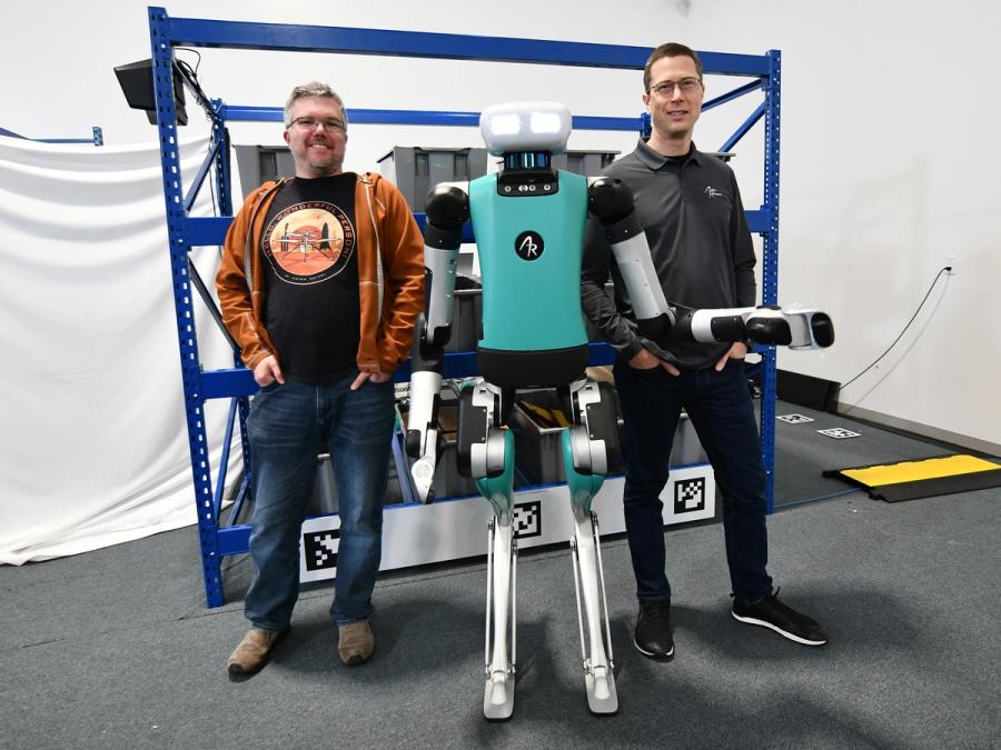 Two human men in casual clothes stand with a Digit humanoid robot that is almost their height.