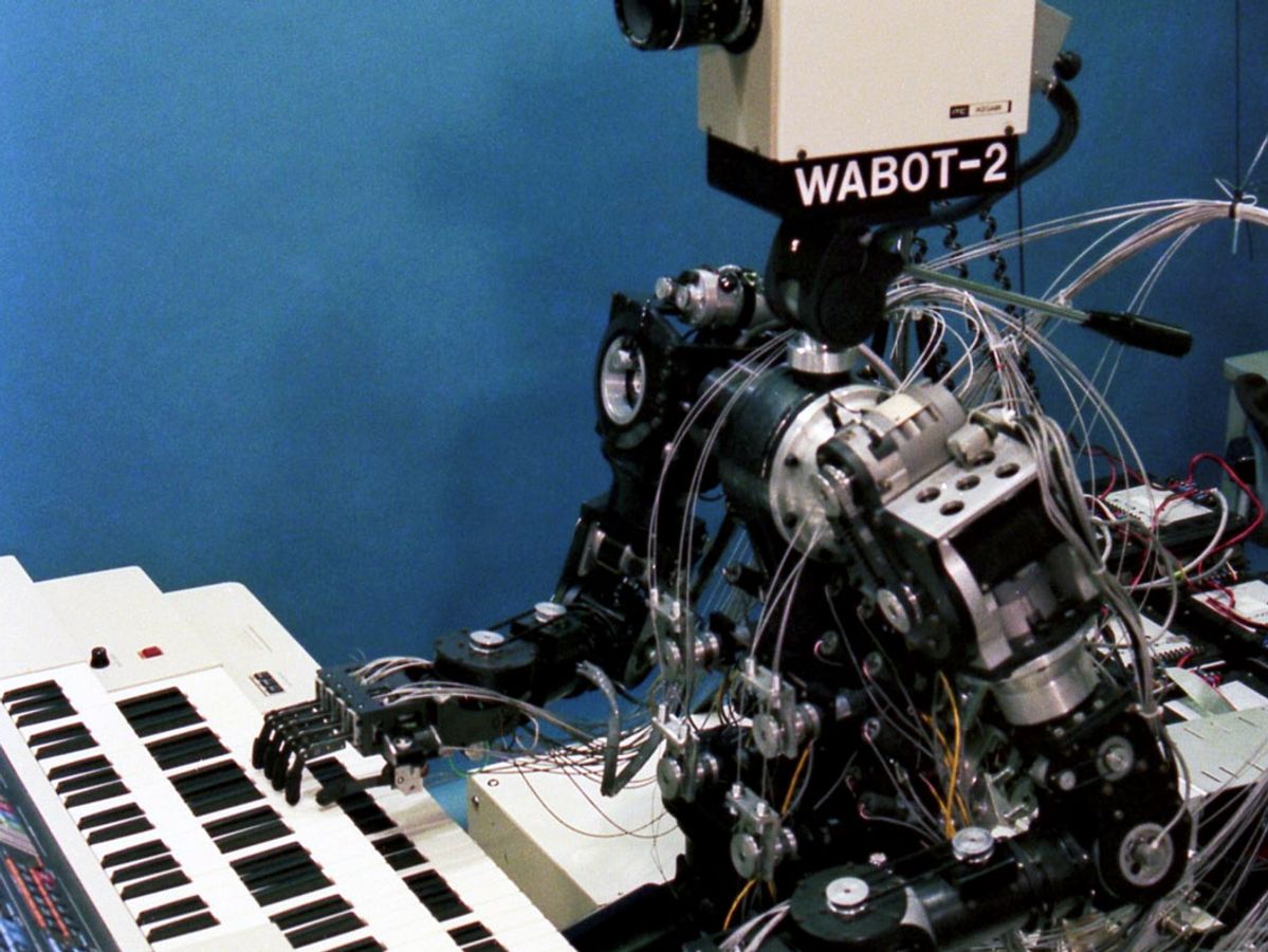 A historical robot with a complex humanoid body and a 1980's era video camera for a head sits and plays the keyboards.