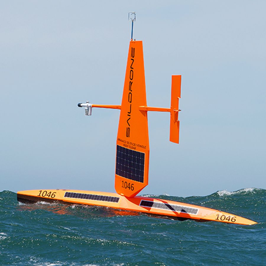 An orange uncrewed surface vehicle consisting of a horizontal unit on the water, with a large sail with solar panels rising vertically, and a rudder running parallel to the sea.