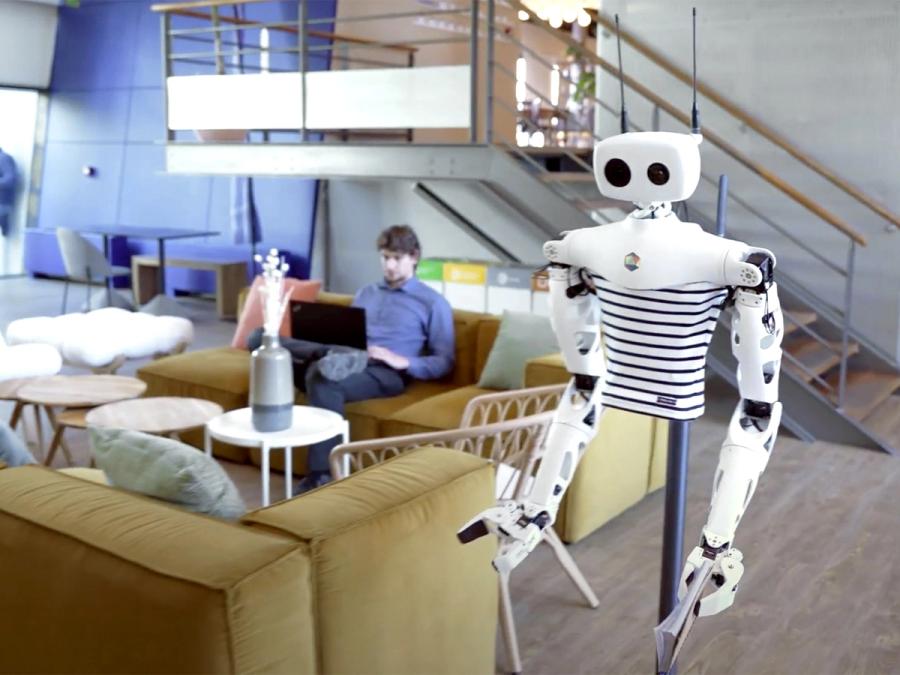 A black and white robotic torso with two white arms ending in two finger gripper hands, and a white head with two antenna, and two camera eyes, one large and one small moves through an office where someone is sitting with a laptop on a couch.