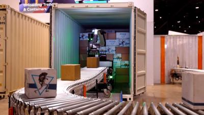 Stretch, a large heavy robot with a mobile wheeled base and a big articulated manipulator arm with suction cups, removes cardboard boxes from inside a white-colored metal contained and places them onto a conveyor belt at a logistics trade show.