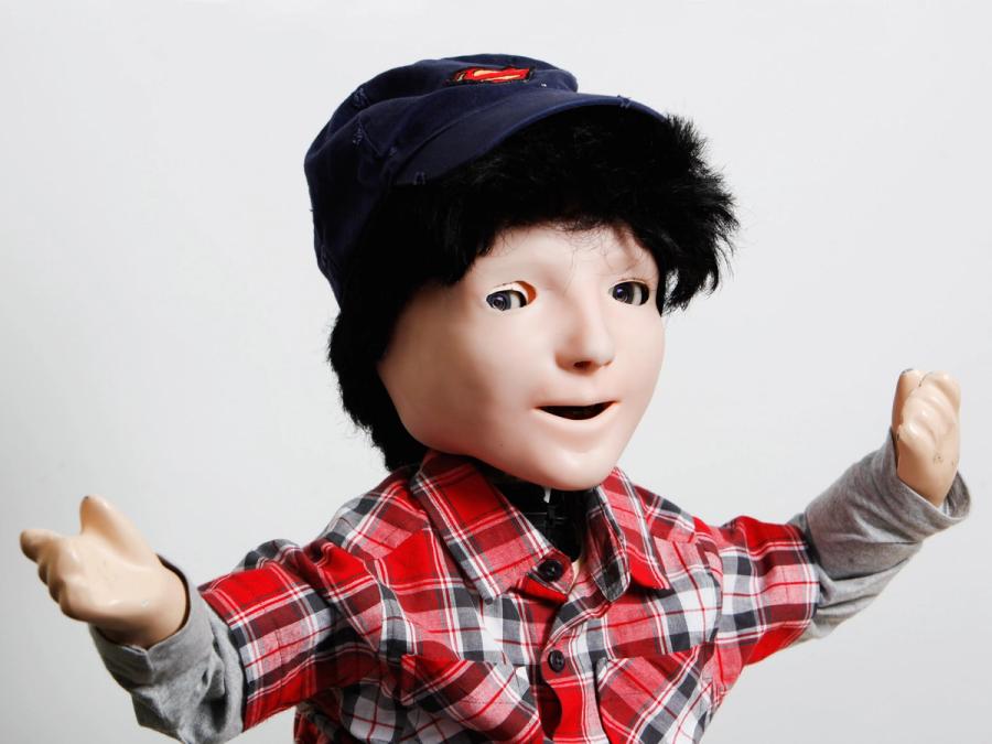 A child-size humanoid with a peachy flesh-like face, eyes, nose and open mouth. It wears a black wig, blue cap and plaid shirt and extends its short arms out.