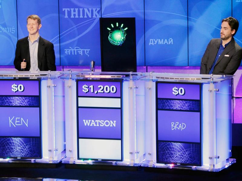 Two people and a vertical display with the IBM Watson logo stand behind attached podiums with screens that read dollar amounts and each individual's name. Watson has $1,200 while Ken and Brad have $0.