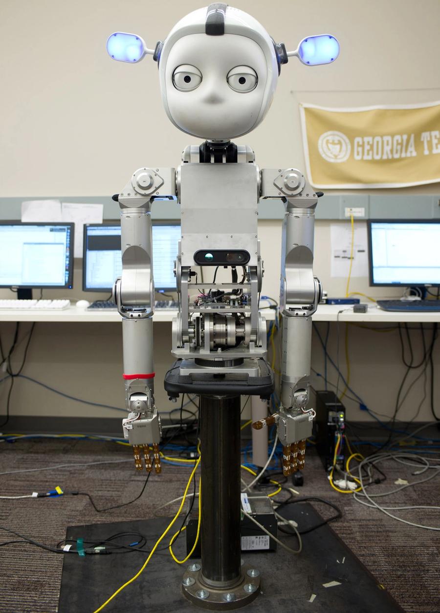 A robot with a round white face with two big eyes, large protruding ears that glow blue, and a silver torso, hands and arms is on a black stand in a lab.