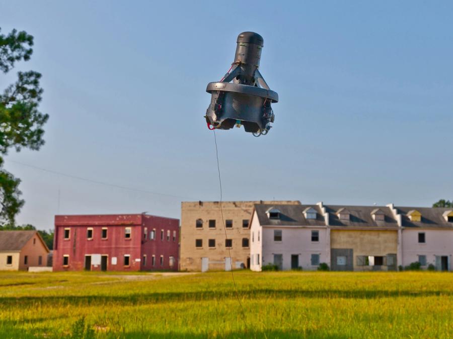 A flying 40 x 30 cm black robot consisting of a base and tall round protrusion. In the background is a row of houses and buildings.