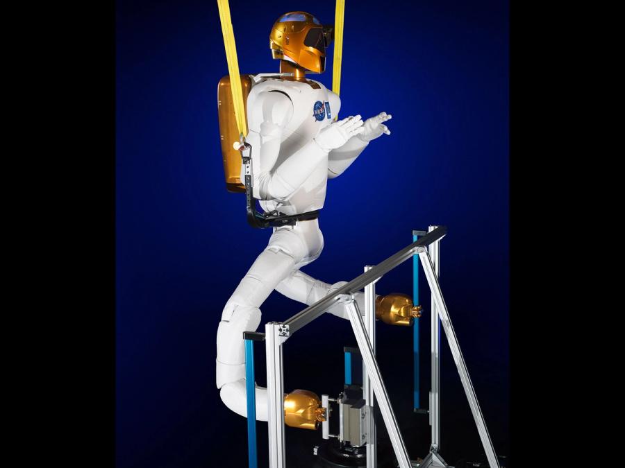 The robot is suspended in a harness. Two soft jointed white legs with golden end effectors are attached to its torso. They manipulate objects below.