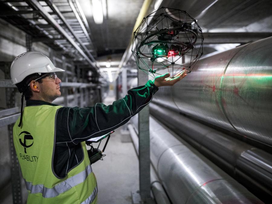 A worker wearing safety clothing and helmet holds the drone towards a pipe.