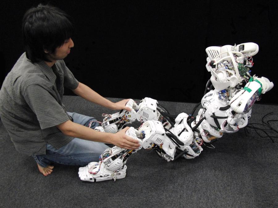 A crouched man holds the legs of a humanoid robot as it does a sit up.