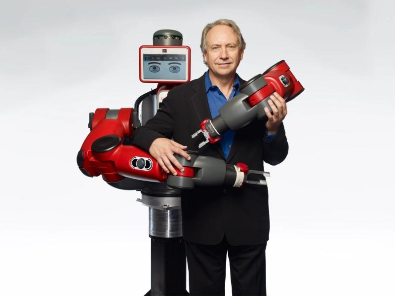 Baxter, a human sized red robot wraps its arms around Rodney Brooks, a smiling man in a suit.