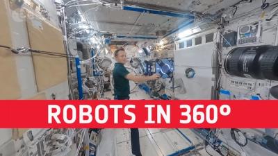 A male astronaut floats inside the space station while gently touching a blue robot cube that also floats, and in front of them a red banner has the words ROBOTS IN 360 DEGREES.