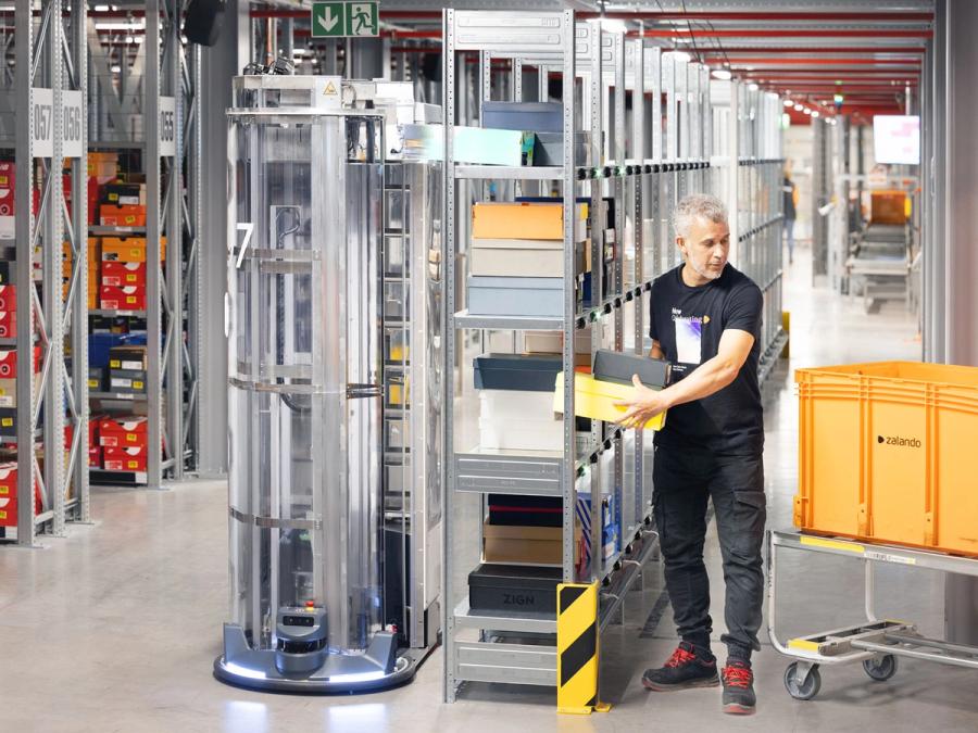 A man moves shoe boxes in a warehouse from shelves to a cart while a tall mobile robot deposits shoe boxes onto the shelves.