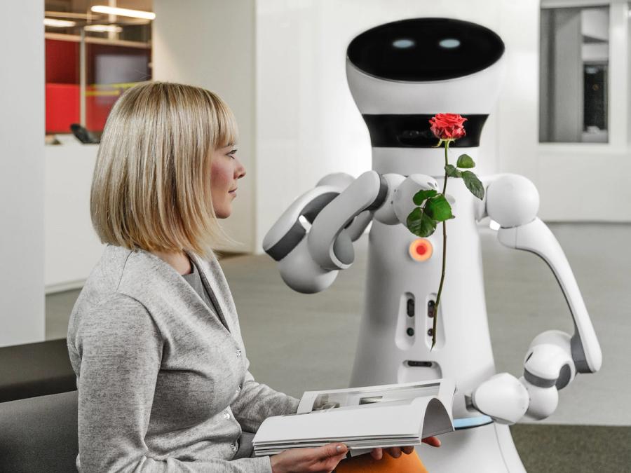 The robot holds a flower out to a seated woman.