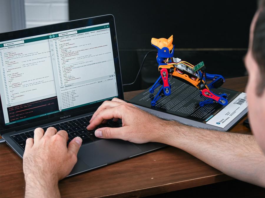 A person codes at a computer, which is attached by a wire to a miniature robot dog.