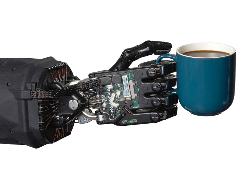 A black prosthetic hand that imitates human fingers and tendons holds a mug of a dark liquid.