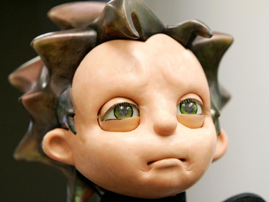 Close-up of humanoid robot with an expressive face, green eyes, flesh-like skin, and brown spiky hair