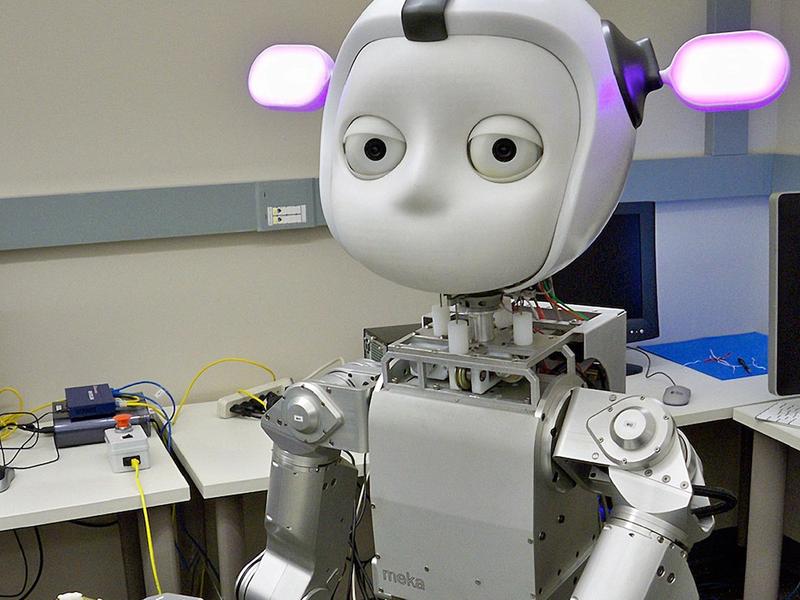 A robot with a metal body, round face with big eyes and pink ears, holds a blue object and looks at the camera, with a lab in the background.