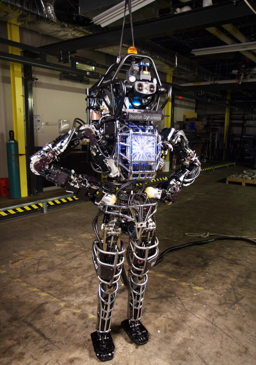 A two legged robot with its hands on hips poses in a warehouse.
