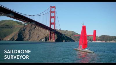 A Saildrone Surveyor, which looks like a large boat with red sail, travels on calm blue water near the Golden Gate Bridge in San Francisco.
