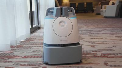 A gray and white plastic robotic vacuum slightly bigger than an upright vacuum drives itself down a carpeted hotel hallway.