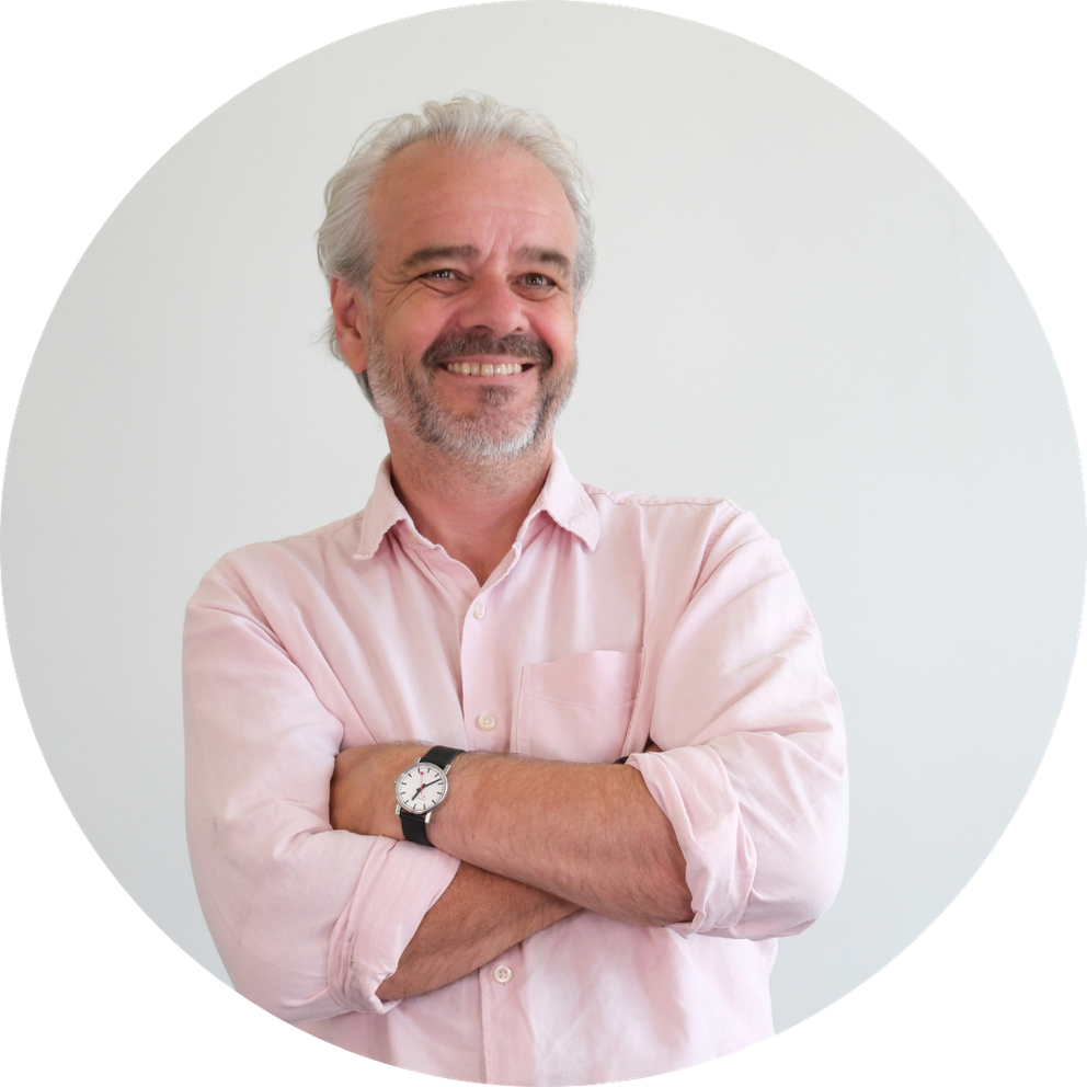 Man wearing a pink shirt stands with arms folded smiling