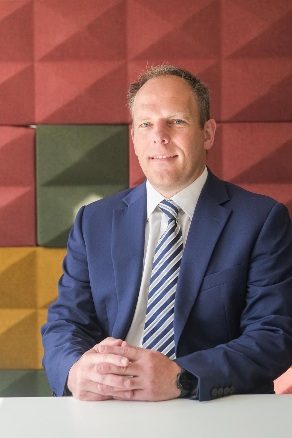 man wearing a suit and tie sitting at a desk and smiling at the camera