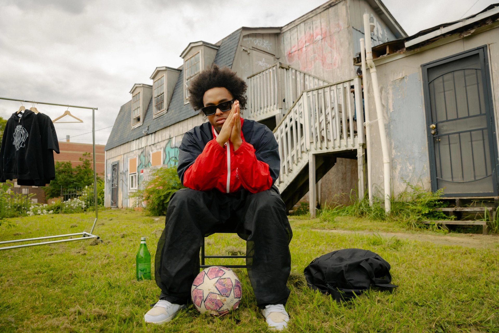 Sajaad seated outdoors in a red and black jacket, wearing sunglasses with his palms together.