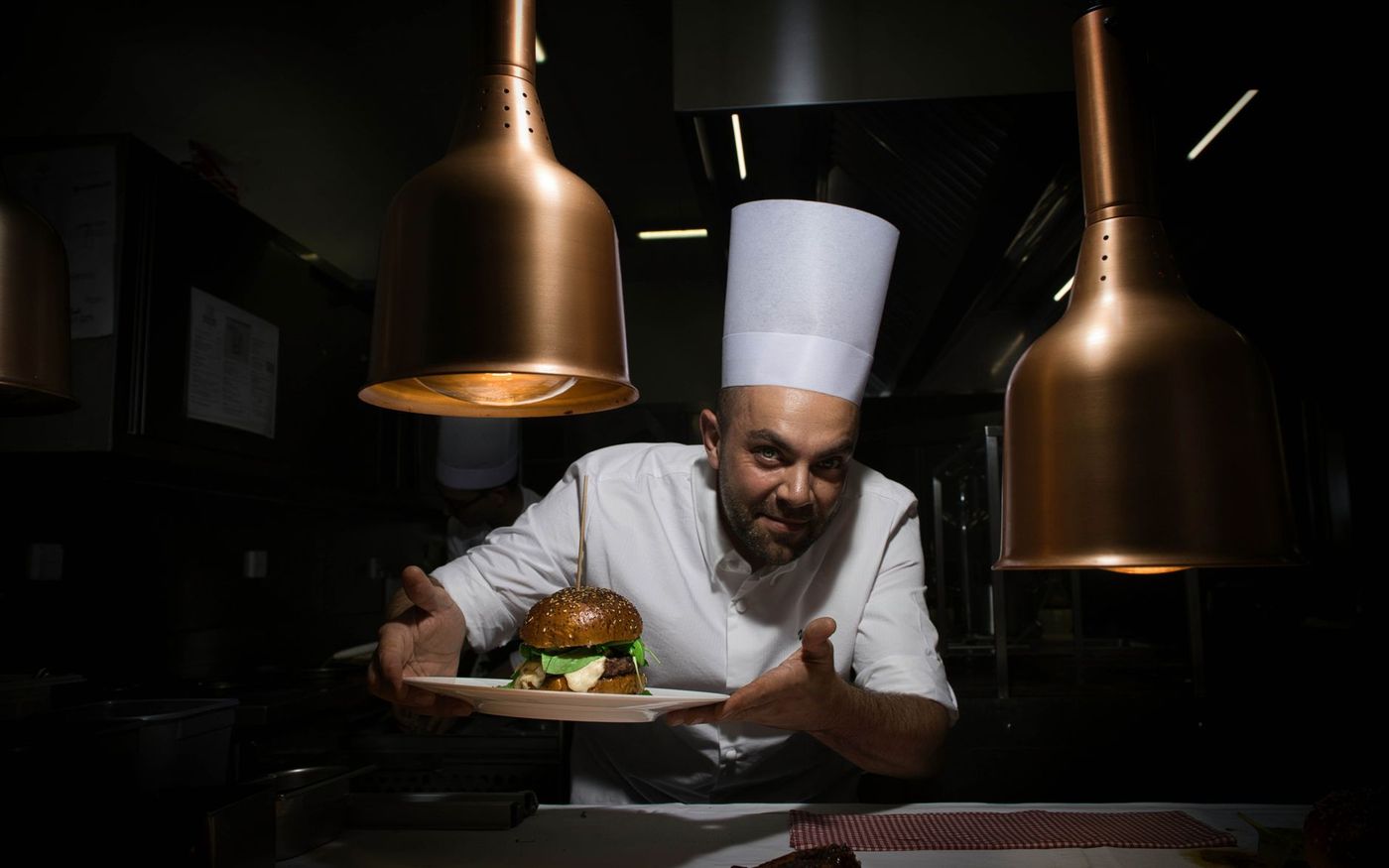 Chef finaliza um hambúrguer em restaurante contemporâneo, com clientes ao fundo. — Foto: Alexandr Zhukovskyi (Pexels)