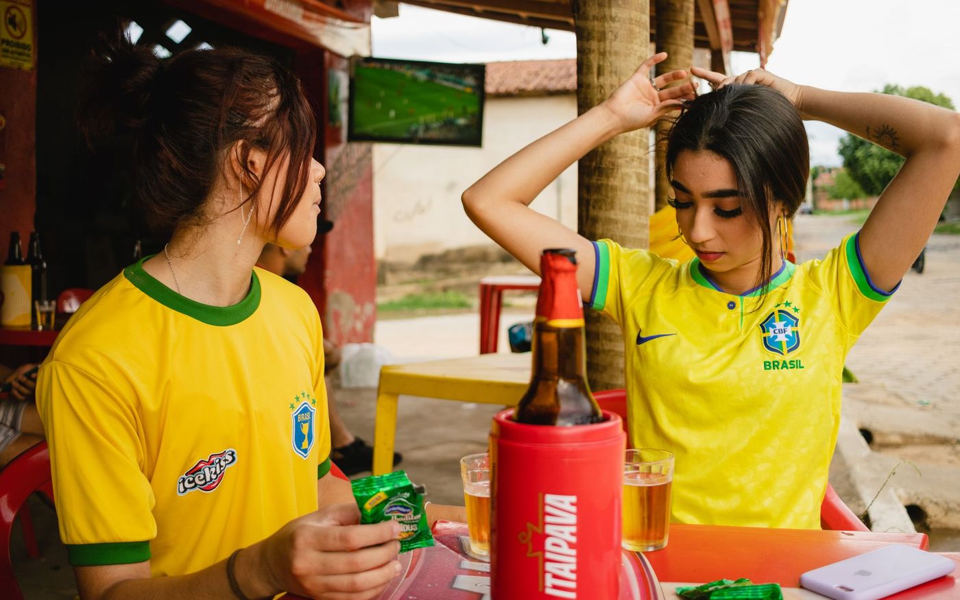 Bar lotado durante transmissão de jogo da Copa do Mundo, clientes torcendo e bebendo drinques coloridos.