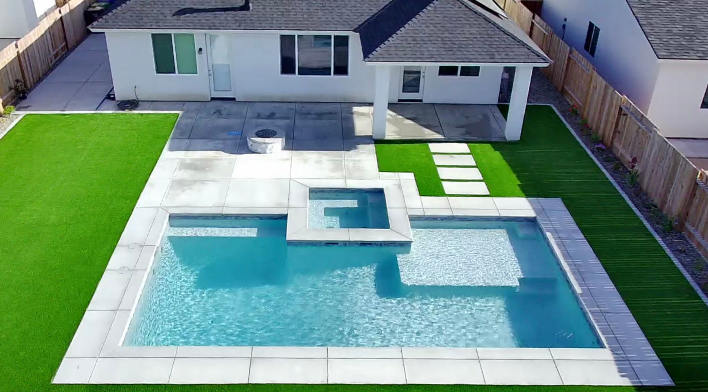 Luxury modern pool with spa, firepit, white concrete pavers, lush artificial turf, and elegant stepping stone pathway 