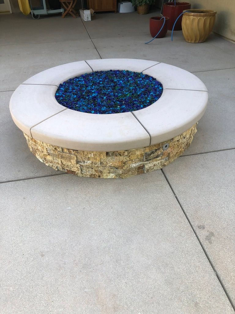 Round gas fire pit with white concrete cap, stacked stone veneer sides, and blue fire glass on smooth concrete patio