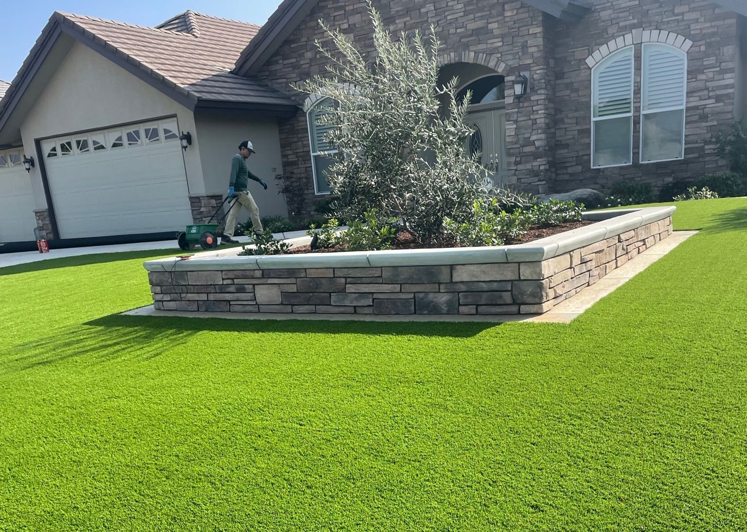 Bright green artificial turf and a raised garden bed made of stacked stone with plants and a small tree in the center