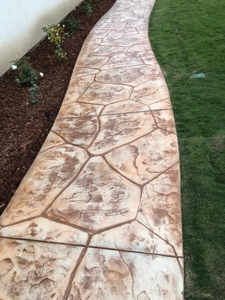 Curved stamped concrete walkway featuring flagstone pattern in tan tones with artificial turf and landscape borders