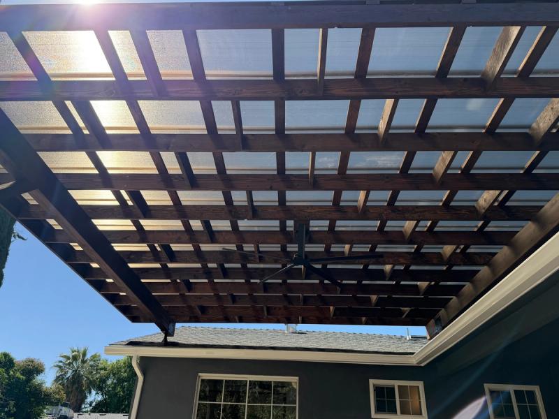 Custom stained wood patio cover with translucent roof panels and integrated ceiling fan, attached to a modern home backyard.