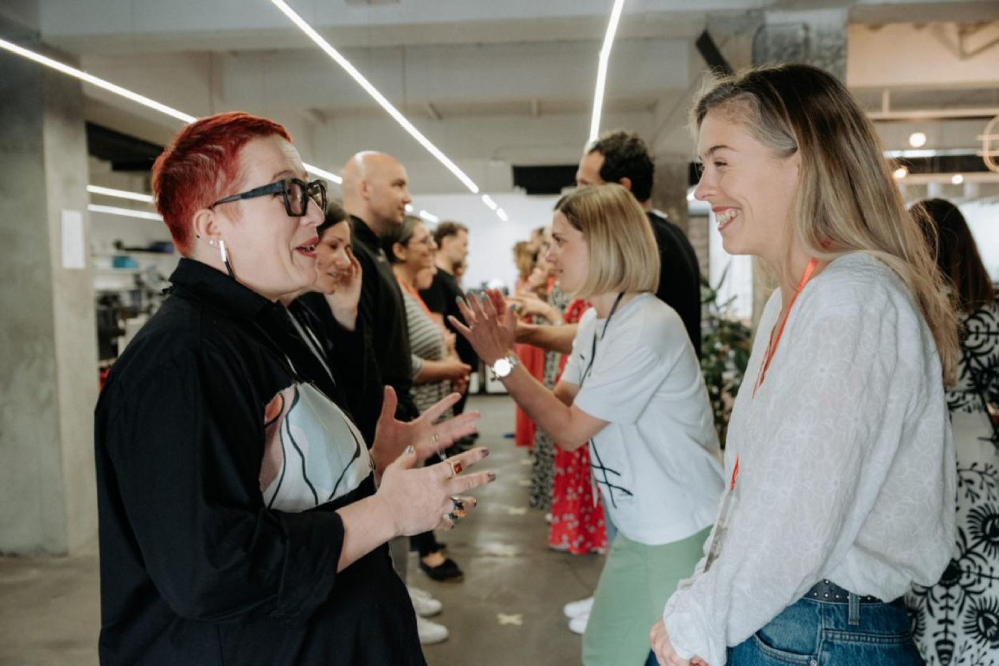Two women smiling and gesturing in the foreground while a group of people interact and clap in the background.