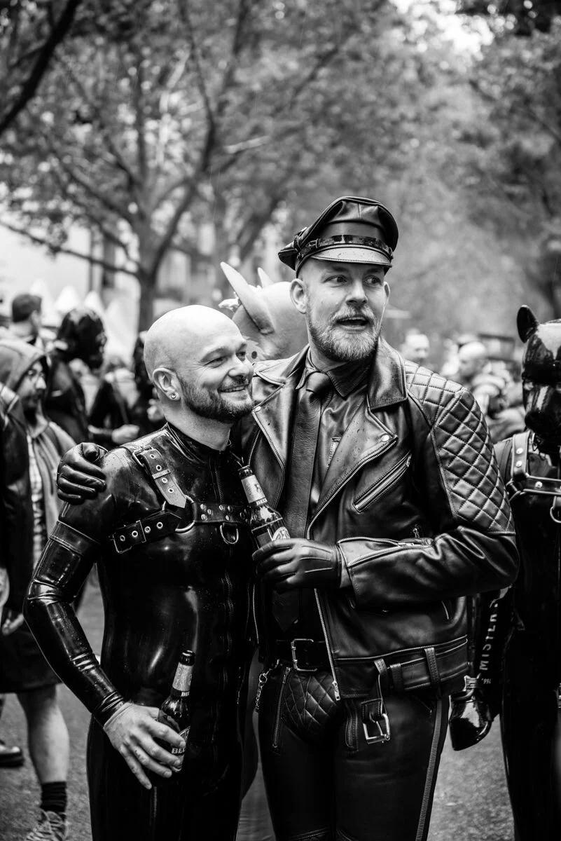 A high-contrast black and white photograph of two men at an outdoor event, one wearing a full latex suit and harness, the other in a quilted leather jacket and peaked cap, embodying the aesthetic of modern leather and latex subcultures.