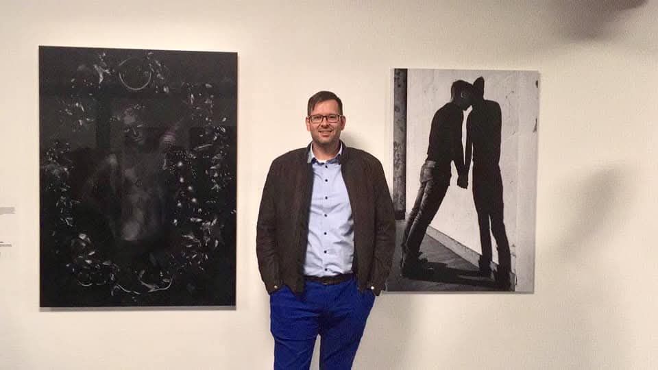 Arjan Spannenburg stands at the Stedelijk Museum Schiedam during the Voor Jillis exhibition, positioned between Erwin Olaf’s dark floral portrait from the Black series and his own vertical black and white photograph titled RELIANT, which depicts two silhouetted figures leaning against a wall.
